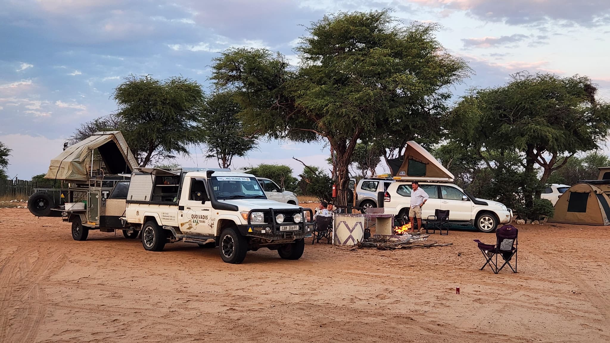 Quo Vadis 4x4 vehicles with rooftop tents pitched around a campfire under camel thorn trees at dusk in the Kalahari