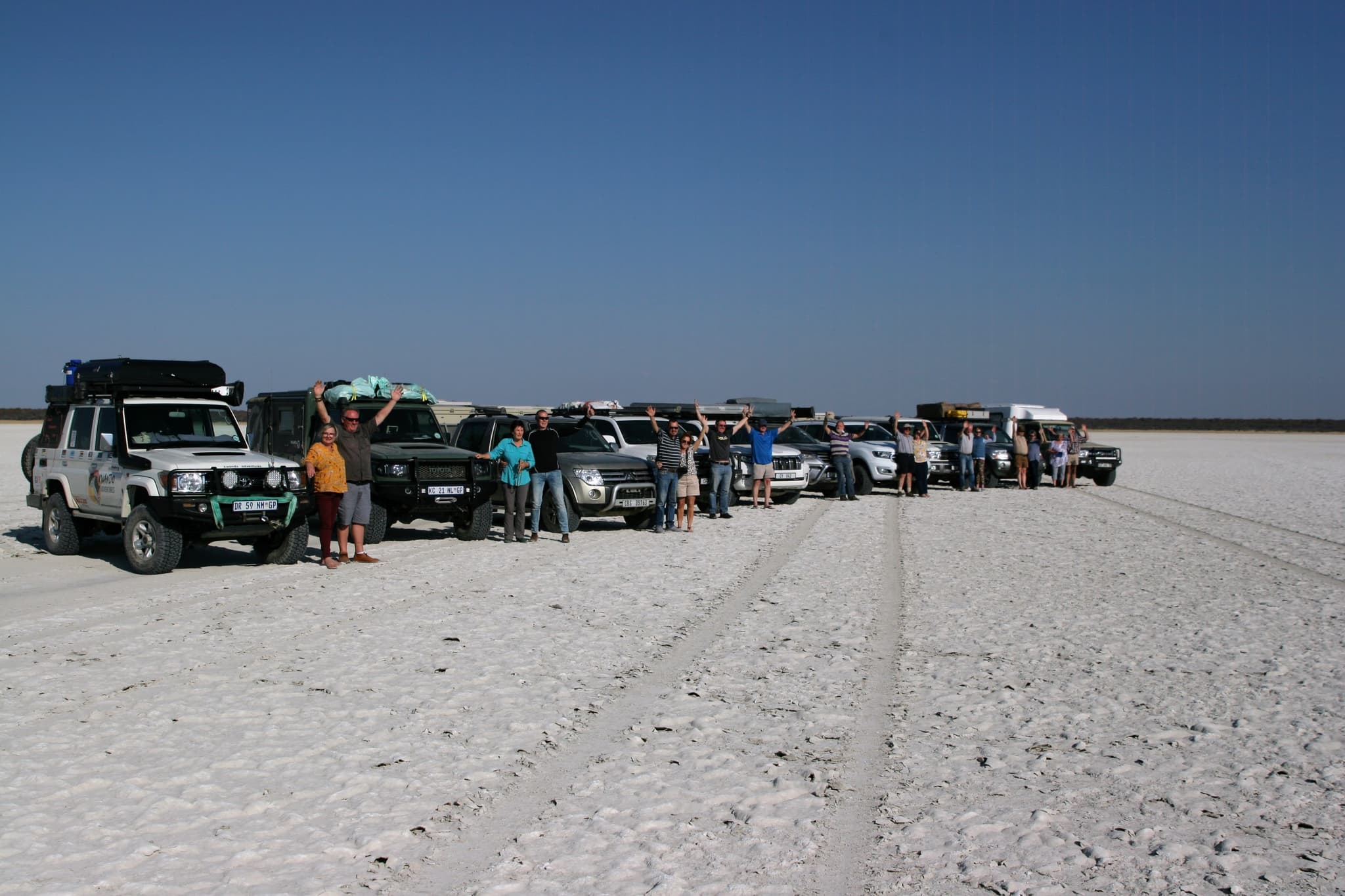 A convoy of guided self-drive 4x4 vehicles lined up on the bright white Makgadikgadi salt pans in Botswana, with travellers waving beside their vehicles under a deep blue sky