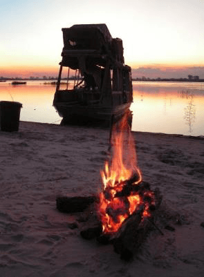 Campfire burning on a sandy riverbank at sunset with a moored houseboat silhouetted against the pink-orange sky