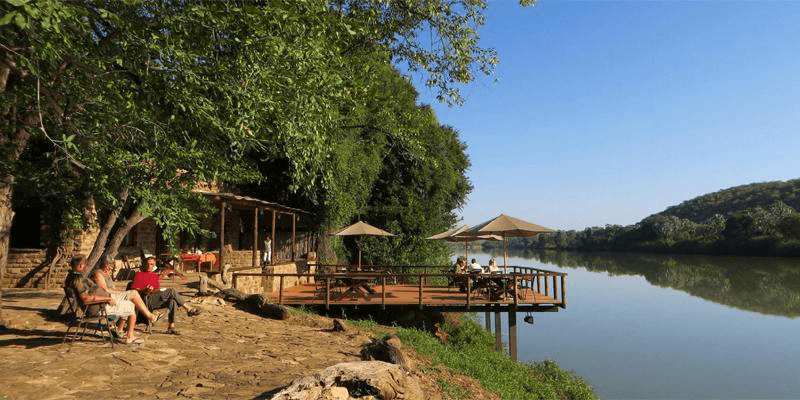 Riverside stone terrace with thatched umbrellas and travellers relaxing in chairs under shady trees beside the calm Kunene River with green hills reflected in the water