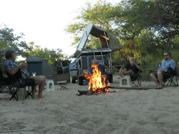 Group of travellers relaxing in camp chairs around a crackling campfire on soft sand at dusk with a 4x4 rooftop-tent vehicle and dome tent among the bushes behind them
