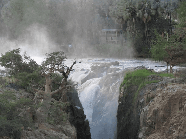 Powerful waterfall plunging over a rocky gorge with mist rising into the air, framed by baobab and makalani palm trees and a small viewing deck