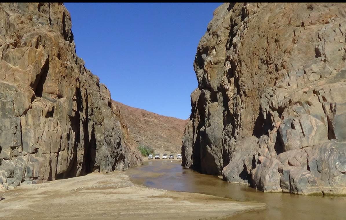 Convoy of 4x4 vehicles driving through a dramatic narrow rocky gorge with steep rugged rock walls towering on both sides beneath a clear blue sky