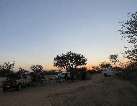 Bush camp at dusk with a Land Rover, rooftop tent and travellers relaxing under an acacia tree as the sun sets over the Botswana bush