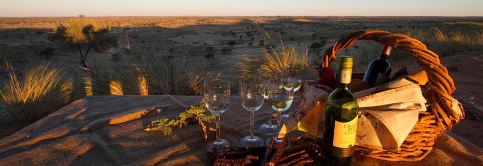 Sundowner-Picknick mit Weingläsern und Korb auf roten Kalahari-Dünen bei Sonnenuntergang