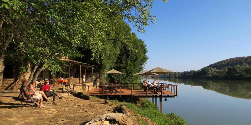 Riverside stone terrace with thatched umbrellas and travellers relaxing in chairs under shady trees beside the calm Kunene River with green hills reflected in the water