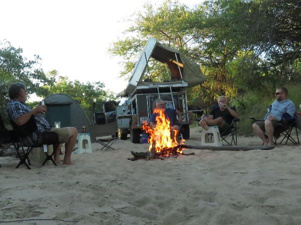 Group of travellers relaxing in camp chairs around a crackling campfire on soft sand at dusk with a 4x4 rooftop-tent vehicle and dome tent among the bushes behind them