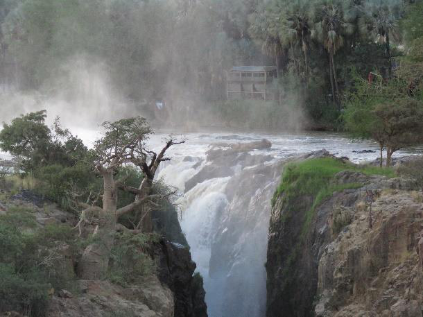 Powerful waterfall plunging over a rocky gorge with mist rising into the air, framed by baobab and makalani palm trees and a small viewing deck