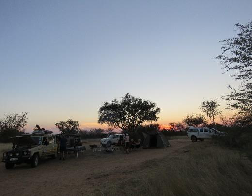 Bush camp at dusk with a Land Rover, rooftop tent and travellers relaxing under an acacia tree as the sun sets over the Botswana bush
