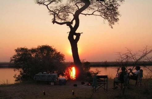 Riverside camp at sunset with a laid dinner table and camp chairs beside a lone tree silhouetted against the sun setting over the Chobe River